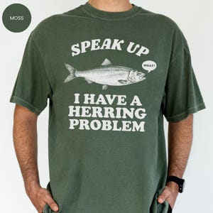 以下が含まれることがあります： モスグリーンのTシャツ。テキスト「SPEAK UP I HAVE A HERRING PROBLEM」と魚のイラストが描かれています。魚は「WHAT？」という吹き出しを持っています。Tシャツは単色です。