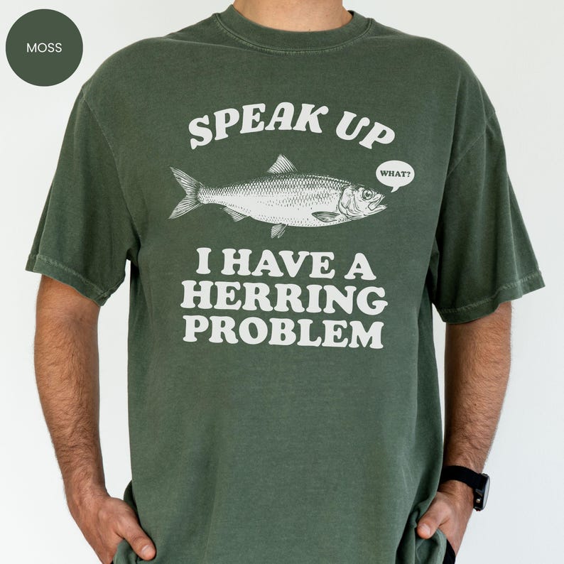 以下が含まれることがあります： モスグリーンのTシャツ。テキスト「SPEAK UP I HAVE A HERRING PROBLEM」と魚のイラストが描かれています。魚は「WHAT？」という吹き出しを持っています。Tシャツは単色です。
