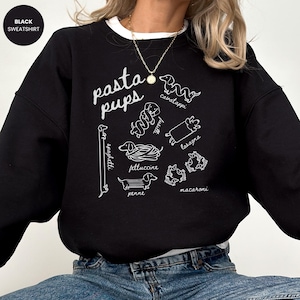 May include: Black sweatshirt with the text "pasta pups" in white script. The design features line drawings of dachshund dogs representing different pasta shapes, including spaghetti, cavatappi, and lasagna. A white crew neck is visible.