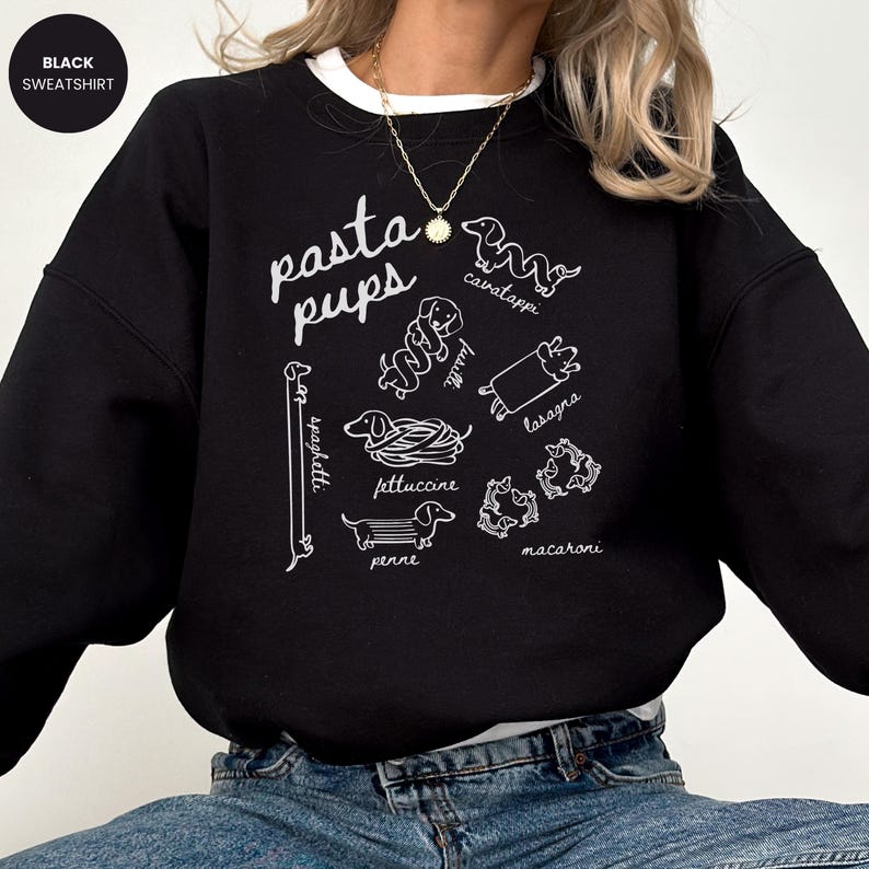 May include: Black sweatshirt with the text "pasta pups" in white script. The design features line drawings of dachshund dogs representing different pasta shapes, including spaghetti, cavatappi, and lasagna. A white crew neck is visible.