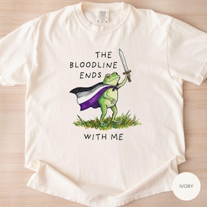 May include: Ivory-colored t-shirt featuring a cartoon frog holding a sword and wearing a cape with the asexual pride flag colors. The text "THE BLOODLINE ENDS WITH ME" is printed above and below the frog illustration.