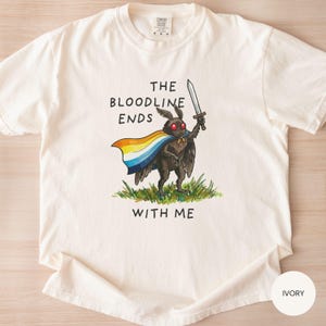 May include: Ivory-colored t-shirt featuring a cartoon moth holding a sword and wearing a rainbow cape, standing on grass. The text "THE BLOODLINE ENDS WITH ME" is printed above and below the moth illustration.