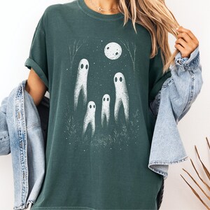 May include: Dark teal t-shirt featuring a white illustration of four ghosts under a full moon. The design includes a night sky with trees and grass. The shirt is a relaxed fit.