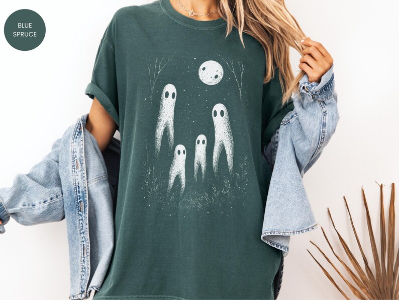 May include: Dark teal t-shirt featuring a white illustration of four ghosts under a full moon. The design includes a night sky with trees and grass. The shirt is a relaxed fit.