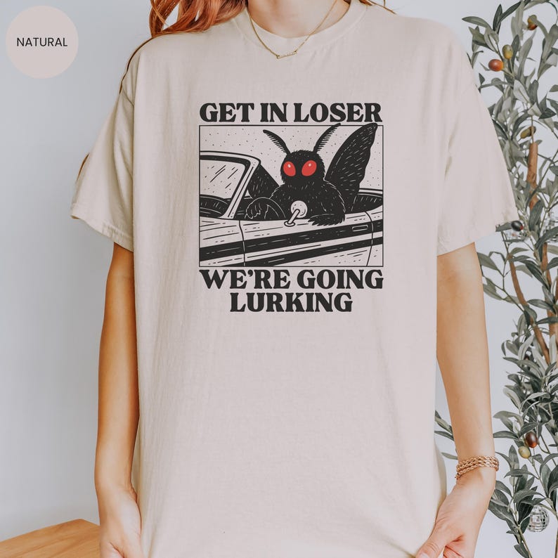 May include: A beige t-shirt featuring a black and white graphic of a moth driving a car. The text "GET IN LOSER" is above the car, and "WE'RE GOING LURKING" is below. The shirt has a crew neck and short sleeves.