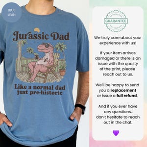 May include: Blue jean t-shirt with a graphic featuring a relaxed dinosaur in sunglasses, the text "Jurassic Dad," and the phrase "Like a normal dad just pre-historic." The design includes palm trees and a wooden deck.