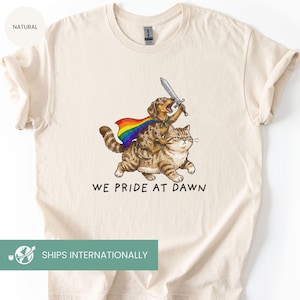 May include: Cream-coloured t-shirt with a playful illustration of a dog riding a cat, both wearing a rainbow cape and holding a sword. The text "WE PRIDE AT DAWN" is printed below the image. The shirt is made of a soft material.