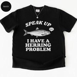 以下が含まれることがあります： 「SPEAK UP I HAVE A HERRING PROBLEM」のテキストと魚のイラストが描かれた黒いTシャツ。魚は「WHAT？」という吹き出しを持っています。このシャツはクルーネックです。