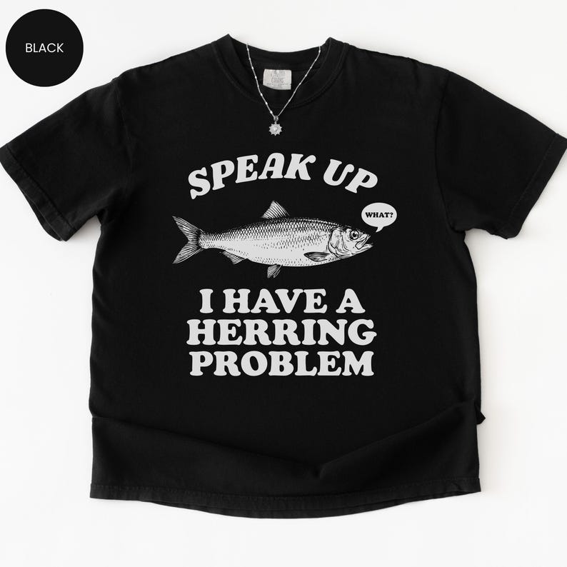 以下が含まれることがあります： 「SPEAK UP I HAVE A HERRING PROBLEM」のテキストと魚のイラストが描かれた黒いTシャツ。魚は「WHAT？」という吹き出しを持っています。このシャツはクルーネックです。