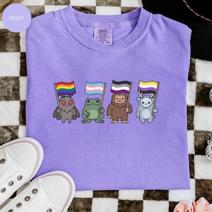 May include: A violet Comfort Colors t-shirt featuring four cartoon creatures, each holding a different pride flag. The creatures include a moth, frog, sasquatch, and a skull-headed deer. The shirt is paired with white sneakers.