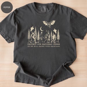 May include: Pepper-colored t-shirt featuring a cream-colored graphic of forest creatures, a bat, and the moon. The text reads, "PROTECT OUR NATIONAL PARKS OR WE WILL HAUNT YOUR BACKYARD."