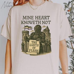May include: A beige t-shirt with a graphic print of a knight holding a scroll that reads "The Lusts of Flesh". The knight is standing in front of a castle with a flag that has a purple, white, and green stripe. The text above the knight reads "Mine Heart Knoweth Not".