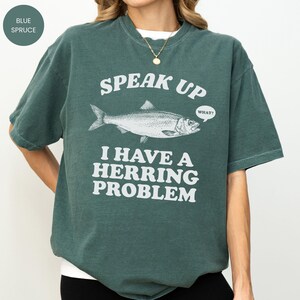 以下が含まれることがあります： 「SPEAK UP」の文字と魚のイラスト、そして「I HAVE A HERRING PROBLEM」というフレーズがプリントされた、ブルー・スプルース色のTシャツ。吹き出しには「WHAT？」と書かれています。