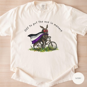 May include: Ivory-colored t-shirt with a graphic of a moth-like creature riding a bicycle, wearing a cape with the asexual pride flag colors. The text "Off to put the ace in menace" is arched above the image. The shirt is a Comfort Colors brand.
