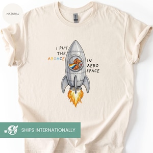 May include: Cream-colored t-shirt featuring a cartoon rocket ship with a dachshund in the window, wearing a cape and holding a glass. The text reads "I PUT THE AROACE IN AERO SPACE."