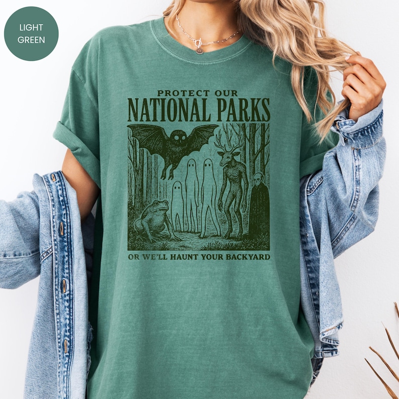 May include: Light green t-shirt with the text "PROTECT OUR NATIONAL PARKS" and the phrase "OR WE'LL HAUNT YOUR BACKYARD". The graphic features a bat, forest creatures, and a frog in a spooky forest scene.
