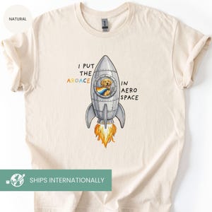 May include: Cream-colored t-shirt featuring a cartoon rocket ship with a golden retriever inside. The rocket has the text "I PUT THE AROACE IN AERO SPACE" in black, orange, and blue. The shirt has a crew neck and short sleeves.
