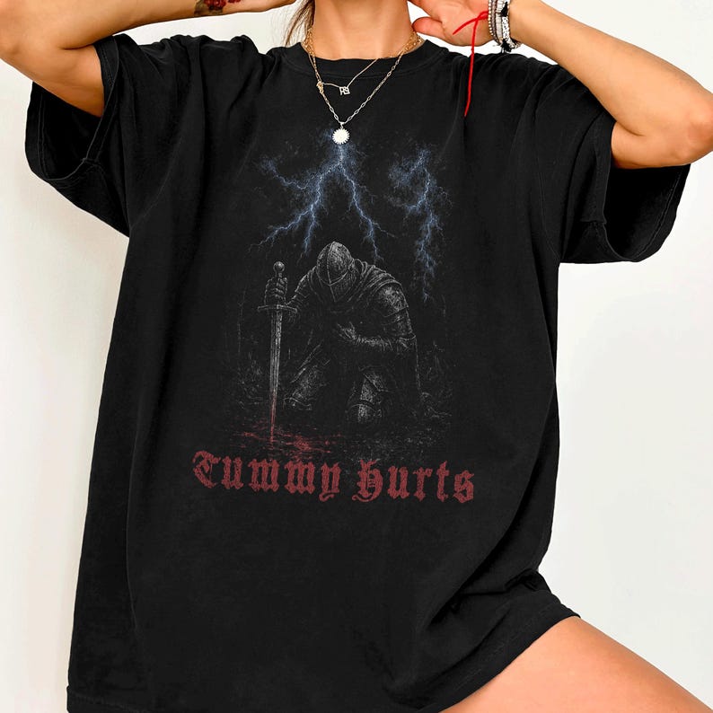 May include: A black t-shirt featuring a graphic of a knight in armor kneeling with a sword, set against a backdrop of blue lightning. Below the knight, the text "Tummy hurts" is printed in a distressed red gothic font on the front of the shirt.