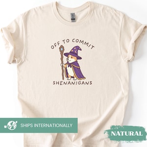Könnte beinhalten: Ein cremefarbenes T-Shirt mit einer Grafik eines Corgis, der als Zauberer mit violettem Hut und Umhang verkleidet ist und einen Holzstab hält. Der Text über und unter der Illustration lautet "OFF TO COMMIT SHENANIGANS" in dunkelbrauner Schrift.