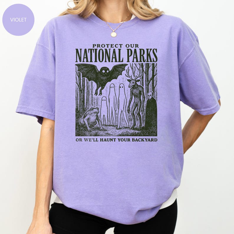 May include: A lilac-coloured t-shirt featuring the text "PROTECT OUR NATIONAL PARKS" and a graphic of forest creatures. The graphic includes a bat, a frog, and several ghost-like figures. The text "OR WE'LL HAUNT YOUR BACKYARD" is below the graphic.