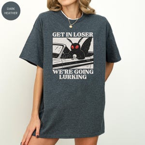 May include: Dark heather gray t-shirt with the text "GET IN LOSER WE'RE GOING LURKING" and a graphic of a moth driving a car. The moth has red eyes and is behind the wheel. The shirt is a casual, oversized fit.