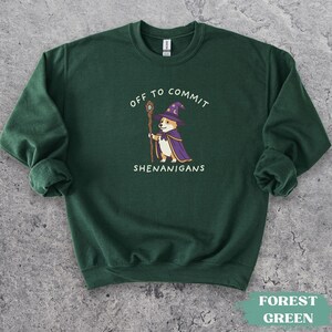 May include: A forest green crewneck sweatshirt featuring a graphic of a corgi dressed as a wizard in a purple hat and cape, holding a wooden staff. The text above and below the illustration reads, "OFF TO COMMIT SHENANIGANS" in white letters.