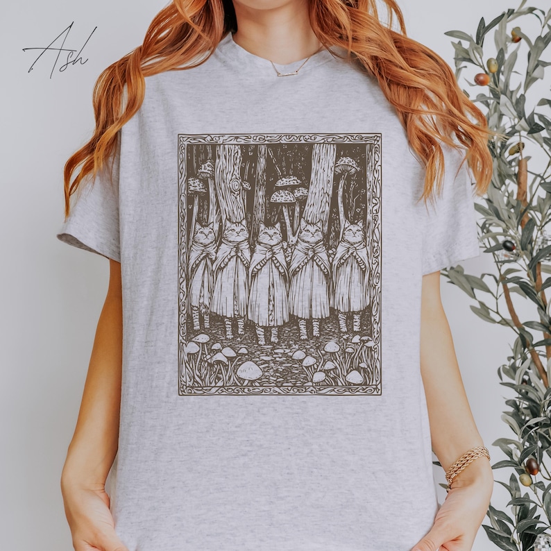 May include: A gray t-shirt with a black and white illustration of five cats in a forest setting. The cats are wearing cloaks and standing in a line. The illustration is framed with a black border.