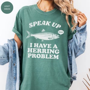 以下が含まれることがあります： 「SPEAK UP I HAVE A HERRING PROBLEM」のテキストと魚のイラストがプリントされた、ライトグリーンのTシャツ。袖はロールアップされ、ゆったりとしたフィット感。テキスト「WHAT？」は吹き出しの中にあります。
