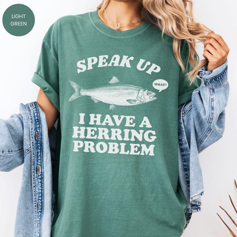 以下が含まれることがあります： 「SPEAK UP I HAVE A HERRING PROBLEM」のテキストと魚のイラストがプリントされた、ライトグリーンのTシャツ。袖はロールアップされ、ゆったりとしたフィット感。テキスト「WHAT？」は吹き出しの中にあります。