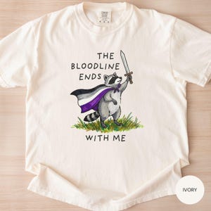 May include: Ivory-colored t-shirt featuring a raccoon illustration. The raccoon, wearing a cape with purple, white, and black colors, holds a sword. Text on the shirt reads "THE BLOODLINE ENDS WITH ME."