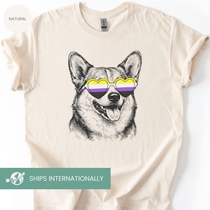 Op de afbeelding: Een crèmekleurig T-shirt met een gedetailleerde illustratie van een Corgi-hond met hartvormige zonnebrillen met gele, paarse en witte glazen. De hond lacht met zijn tong uit. Op het shirt staat het woord "NATURAL".
