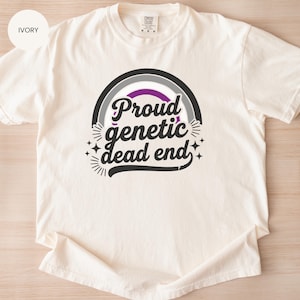 May include: Ivory-colored t-shirt with a crew neck. The front features a graphic with a black and gray arched design, a purple stripe, and the words "Proud genetic dead end" in a stylized font. The design includes starburst and star accents.