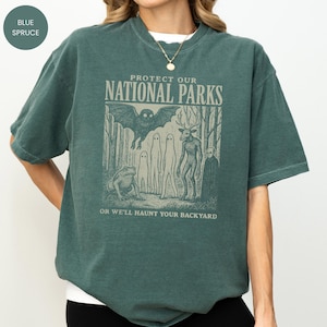 May include: A blue spruce coloured t-shirt with the text "PROTECT OUR NATIONAL PARKS" and the phrase "OR WE'LL HAUNT YOUR BACKYARD". The graphic features a bat, a frog, ghosts, and a deer-like creature in a forest setting.