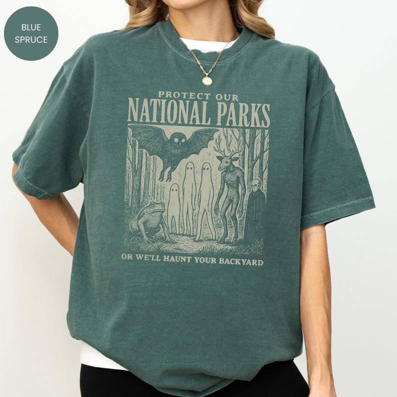 May include: A blue spruce coloured t-shirt with the text "PROTECT OUR NATIONAL PARKS" and the phrase "OR WE'LL HAUNT YOUR BACKYARD". The graphic features a bat, a frog, ghosts, and a deer-like creature in a forest setting.