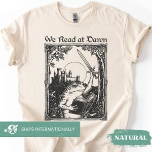 May include: A cream-colored t-shirt featuring a black ink-style illustration of a frog holding a sword and an open book in front of a castle. The text "We Read at Dawn" is printed in a decorative font above the framed graphic design.