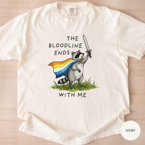 May include: Ivory-colored t-shirt featuring a raccoon illustration. The raccoon, wearing a cape and holding a sword, stands on grass. Text on the shirt reads "THE BLOODLINE ENDS WITH ME."