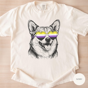 May include: Ivory-colored t-shirt featuring a detailed black and white illustration of a Corgi wearing heart-shaped sunglasses with the non-binary pride flag colors. The shirt has a crew neck and short sleeves. The word "IVORY" is printed on a white circle.