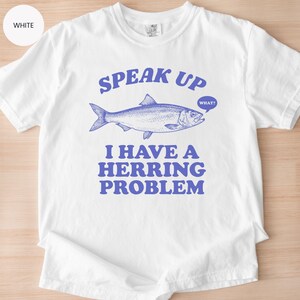 以下が含まれることがあります： 青いニシンのグラフィックと「SPEAK UP」の文字が上にプリントされた白いTシャツ。魚の下には「I HAVE A HERRING PROBLEM」の文字。吹き出しには「WHAT？」と書かれています。