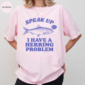以下が含まれることがあります： 薄いピンク色のTシャツ。青いニシンのイラストの上に「SPEAK UP」の文字。魚の下には「I HAVE A HERRING PROBLEM」の文字。吹き出しには「WHAT？」と書かれています。