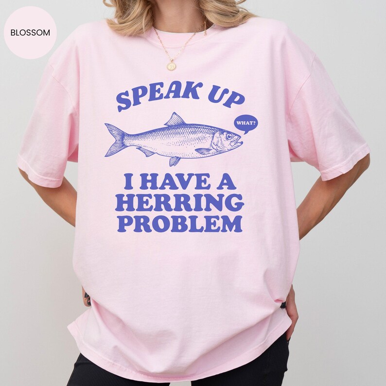 以下が含まれることがあります： 薄いピンク色のTシャツ。青いニシンのイラストの上に「SPEAK UP」の文字。魚の下には「I HAVE A HERRING PROBLEM」の文字。吹き出しには「WHAT？」と書かれています。
