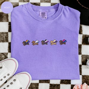 May include: A violet Comfort Colors t-shirt featuring embroidered dachshunds with pride flag capes. The shirt has a crew neck and short sleeves. White sneakers are visible in the bottom left corner.