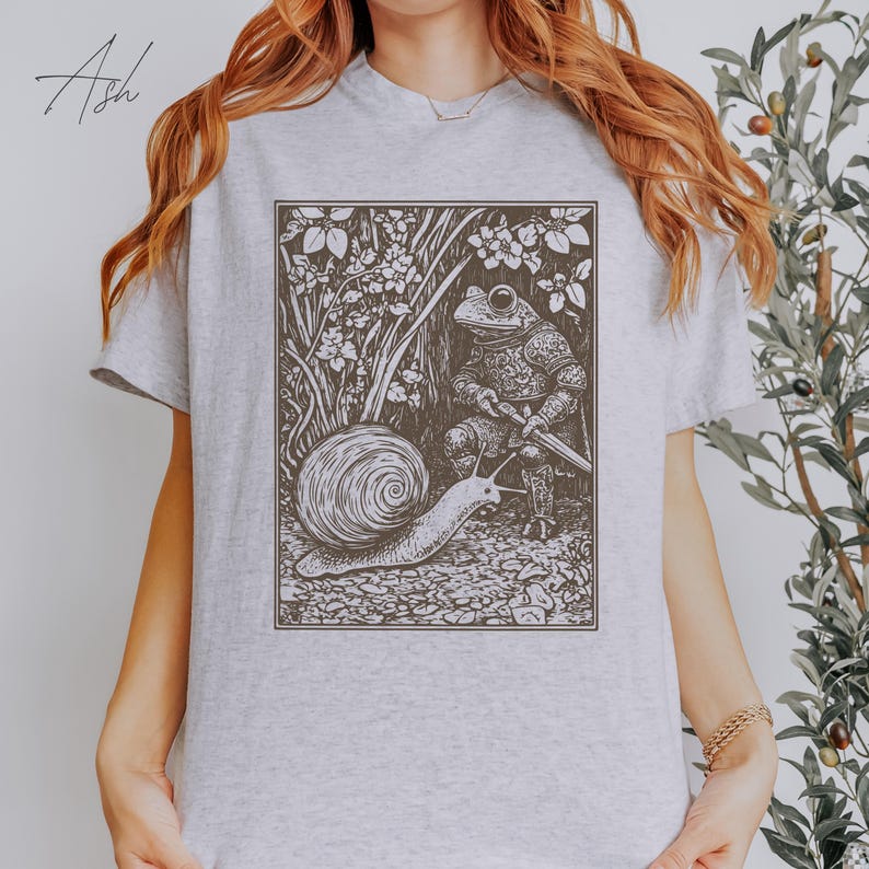K&ouml;nnte beinhalten: Ein graues T-Shirt mit einer Schwarzwei&szlig;illustration eines Frosches in R&uuml;stung, der ein Schwert h&auml;lt, und einer Schnecke, die auf ihn zukriecht.