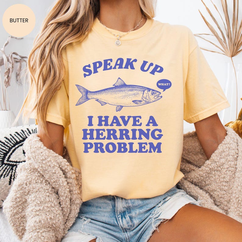 以下が含まれることがあります： バター色のTシャツ。青色の文字で「SPEAK UP I HAVE A HERRING PROBLEM」と書かれ、青い魚のイラストが描かれています。カジュアルでオーバーサイズのフィット感です。