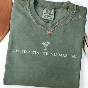May include: A moss green t-shirt with the phrase "I NEED A TINI WEENIE MARTINI" printed in white. A gold necklace with a pendant rests on the shirt. The shirt is folded with a hat and dried plant in the background.
