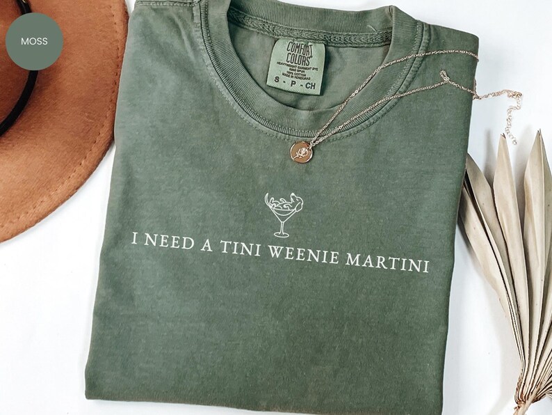 May include: A moss green t-shirt with the phrase "I NEED A TINI WEENIE MARTINI" printed in white. A gold necklace with a pendant rests on the shirt. The shirt is folded with a hat and dried plant in the background.