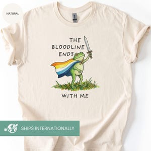 May include: Cream-colored t-shirt featuring a green frog wearing a rainbow cape and holding a sword. The text "THE BLOODLINE ENDS WITH ME" is printed above and below the frog illustration. The shirt is made of a soft material.