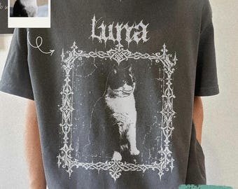 Puede incluir: Una camiseta gris carbón con un estampado gráfico blanco de un gato blanco y negro sentado dentro de un marco rectangular ornamentado. Sobre el marco, el texto "Luna" está impreso en una fuente de estilo gótico. Tiene cuello redondo.