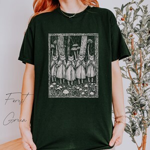 May include: Black t-shirt with a white graphic print of a group of cats wearing cloaks and standing in a forest with mushrooms. The text "Forest Green" is visible in the lower left corner of the image.