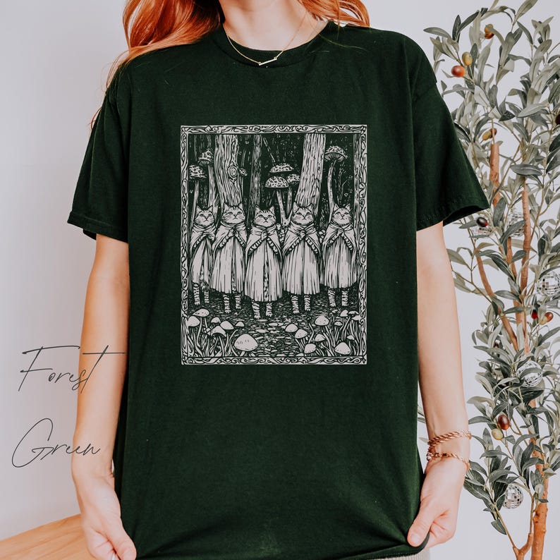 May include: Black t-shirt with a white graphic print of a group of cats wearing cloaks and standing in a forest with mushrooms. The text "Forest Green" is visible in the lower left corner of the image.