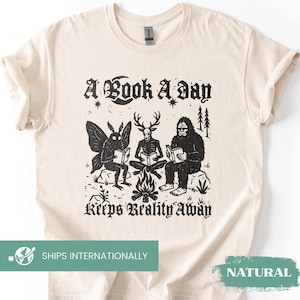 May include: A natural-colored t-shirt featuring a black graphic print of a moth, a deer-headed figure, and Bigfoot sitting around a campfire reading books. The text "A Book A Day Keeps Reality Away" is printed in a gothic font above and below the scene.